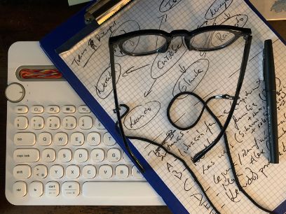 Auf einem schwarzen Hintergrund liegt eine Computertastatur, darüber ein kariertes Blatt Papier und auf dem Papier eine Brille und ein Stift.