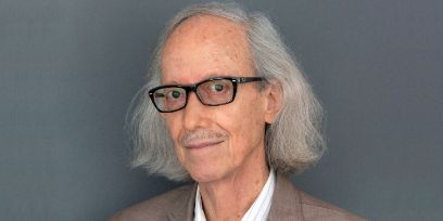 An older man with grey, shoulder-length hair and glasses looks into the camera against a grey background.